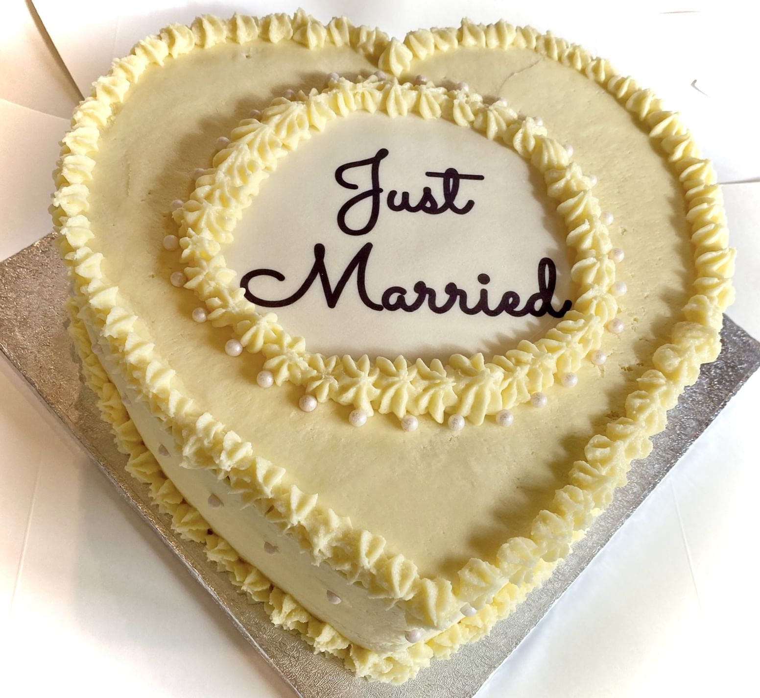 Heart-shaped cake with creamy white frosting. The edges are decorated with piped frosting and small edible pearls. The center of the cake has the words 'Just Married' written in elegant script. The cake is placed on a silver board.